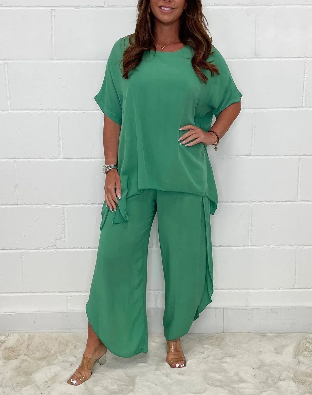 Woman wearing loose green flowy pants and matching top standing against a white brick wall