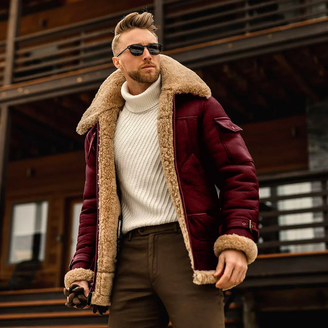 Stylish man wearing burgundy shearling jacket, white turtleneck sweater, and sunglasses outside wooden cabin