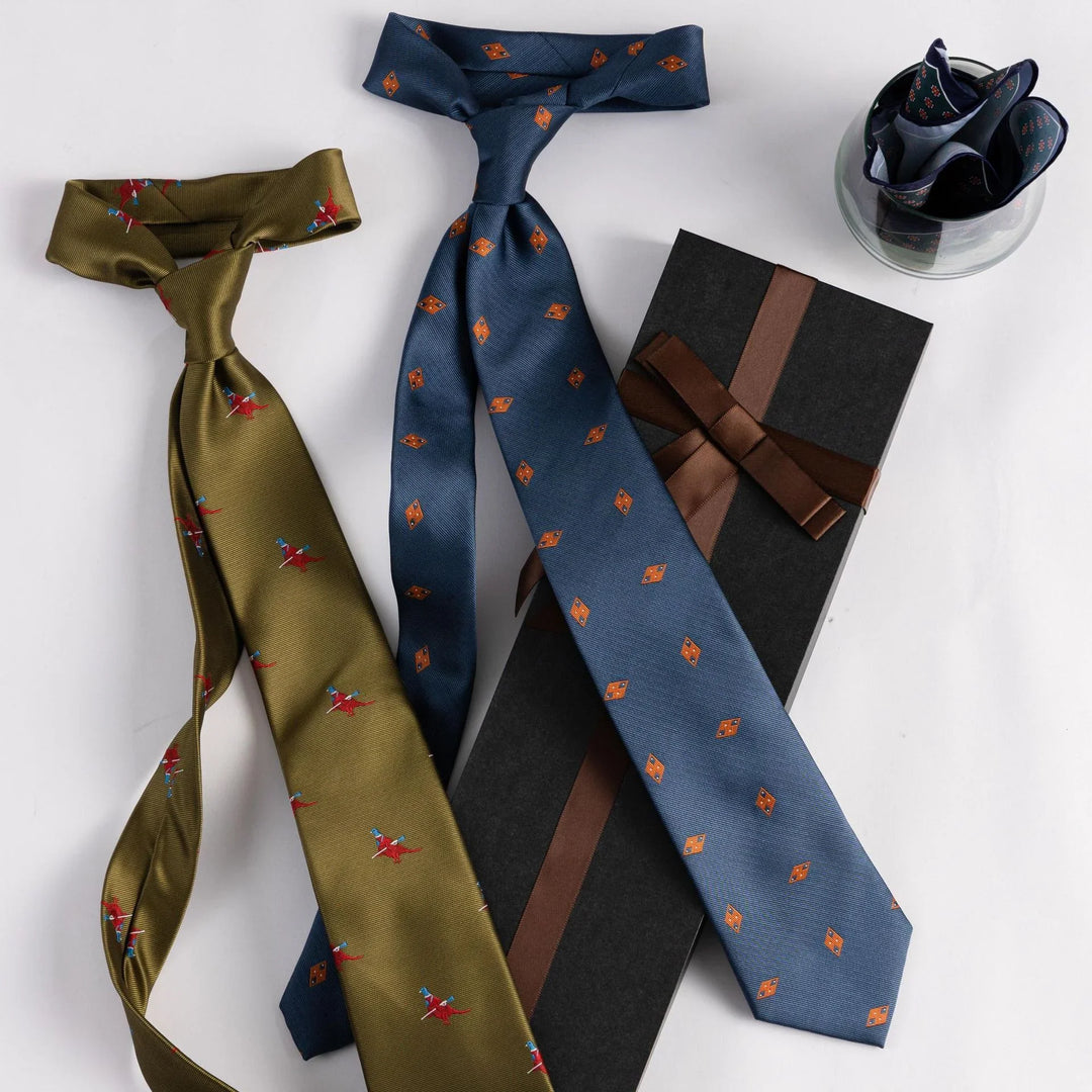 Two patterned silk ties, one olive green with red birds and one blue with orange shapes, next to a black gift box and folded pocket square