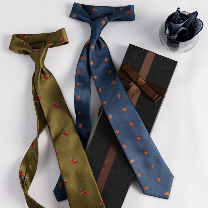 Two patterned silk ties, one olive green with red birds and one blue with orange shapes, next to a black gift box and folded pocket square