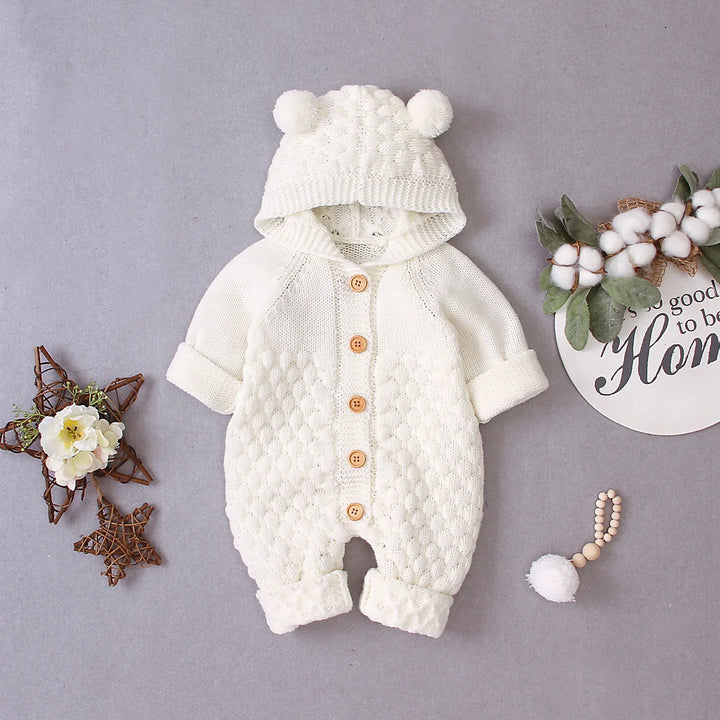 Cream-colored knitted baby romper with hood and pom-poms, wooden buttons, and textured pattern