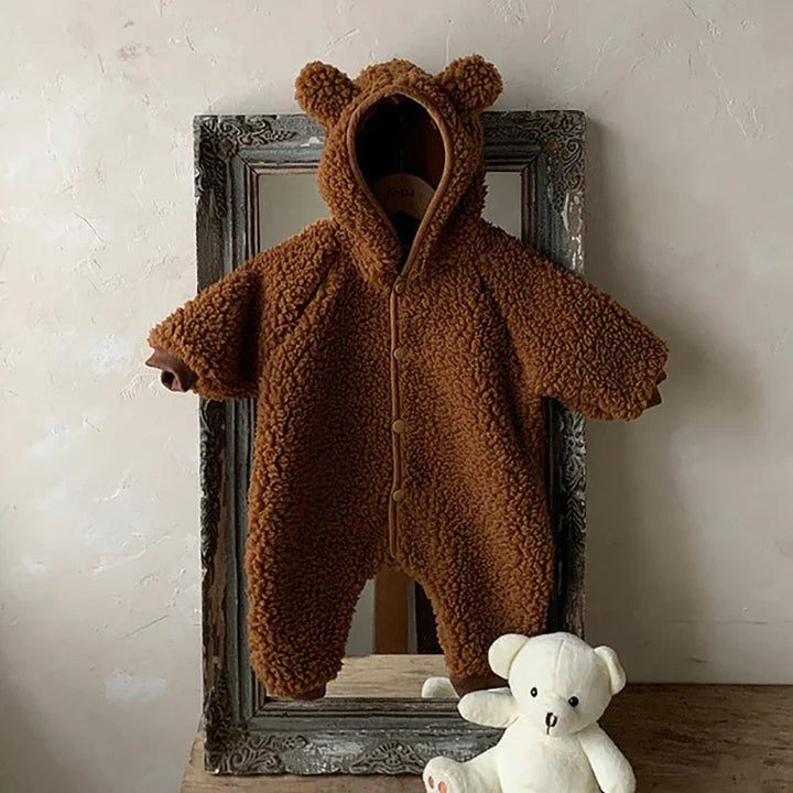 Brown teddy bear fleece baby onesie with hood and ears hanging on vintage frame with white teddy bear plush