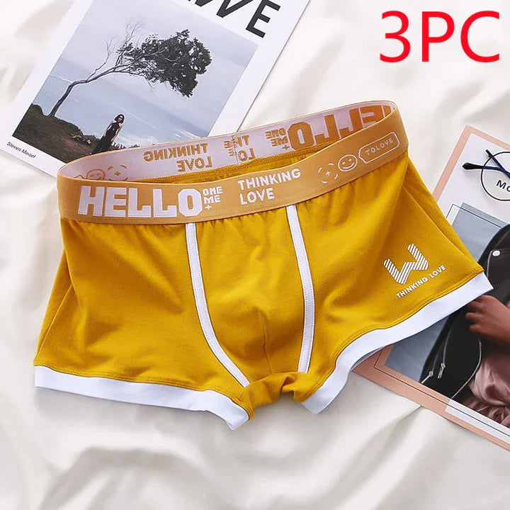 Yellow men's boxer briefs with white trim and branded waistband reading 'HELLO Thinking Love' laid on white fabric with magazines