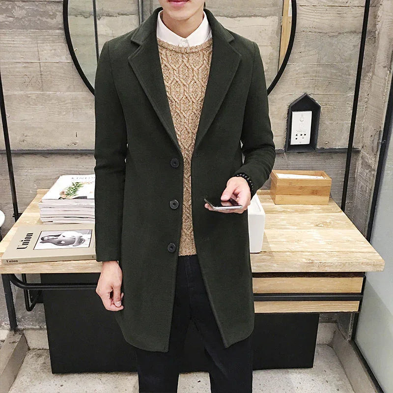 Man wearing dark green wool coat over beige cable knit sweater and white shirt indoors