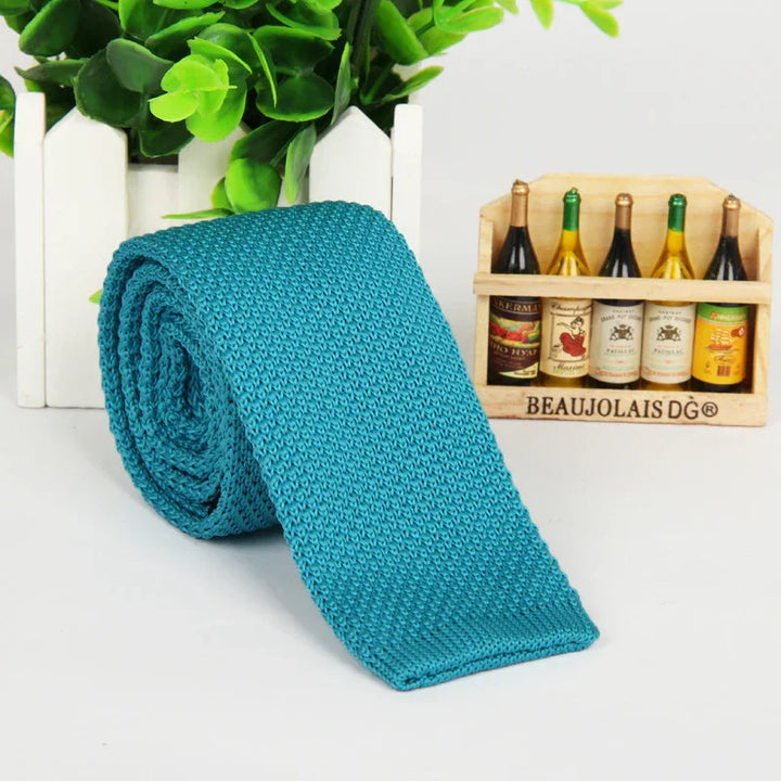 Rolled turquoise knitted tie on white surface with green plant and miniature wine bottle rack in background