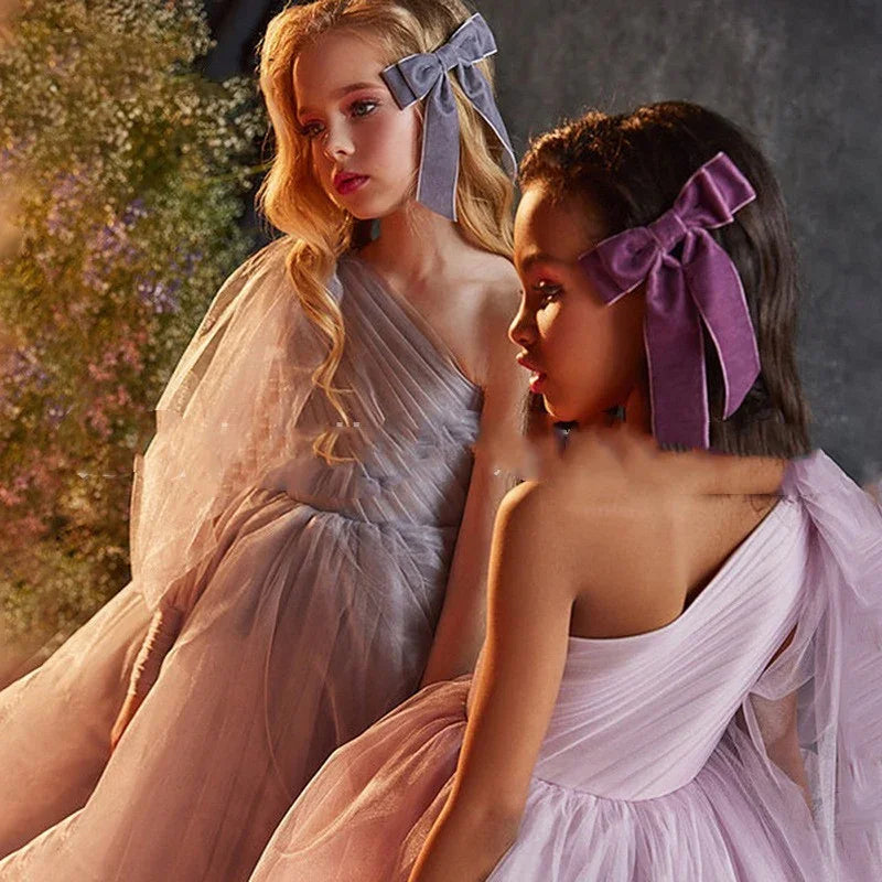 Two young girls in elegant pastel tulle dresses with large velvet hair bows posing in a soft, floral setting