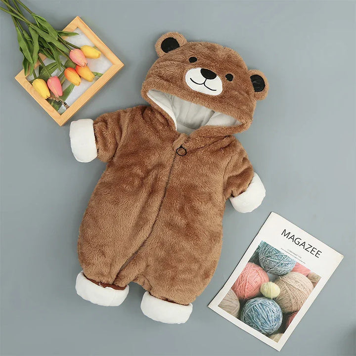 Brown fuzzy baby onesie with bear face hood, white cuffs, lying on gray surface with flowers and yarn magazine