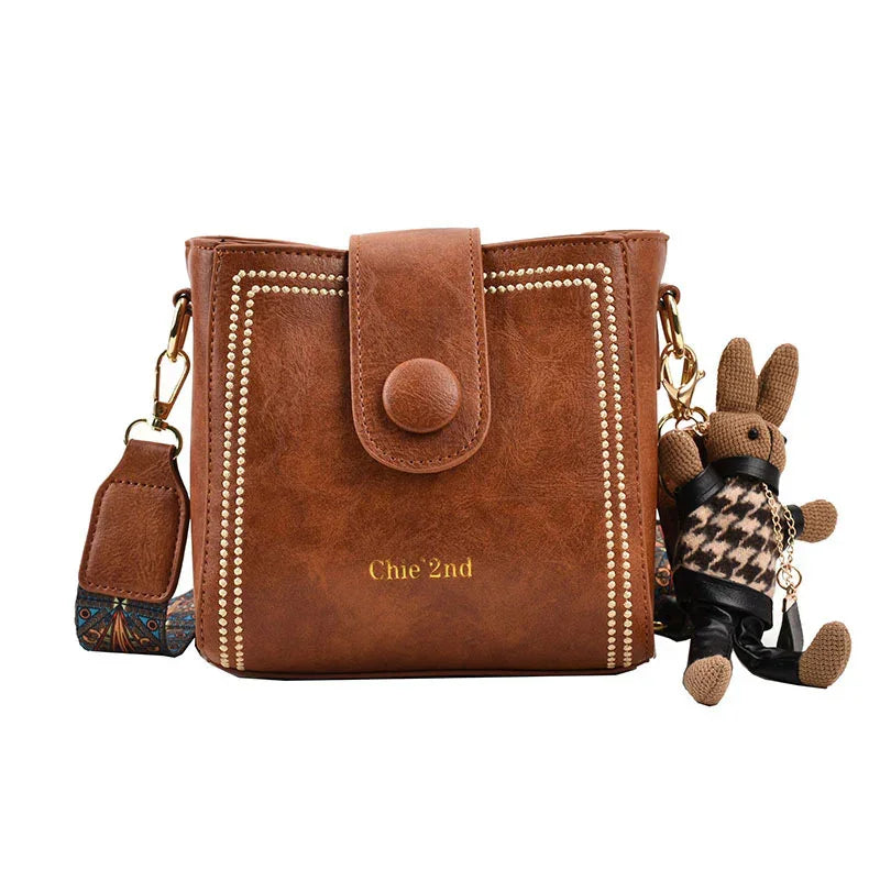 Brown leather crossbody bag with studded border, button flap, patterned strap, and attached bunny keychain