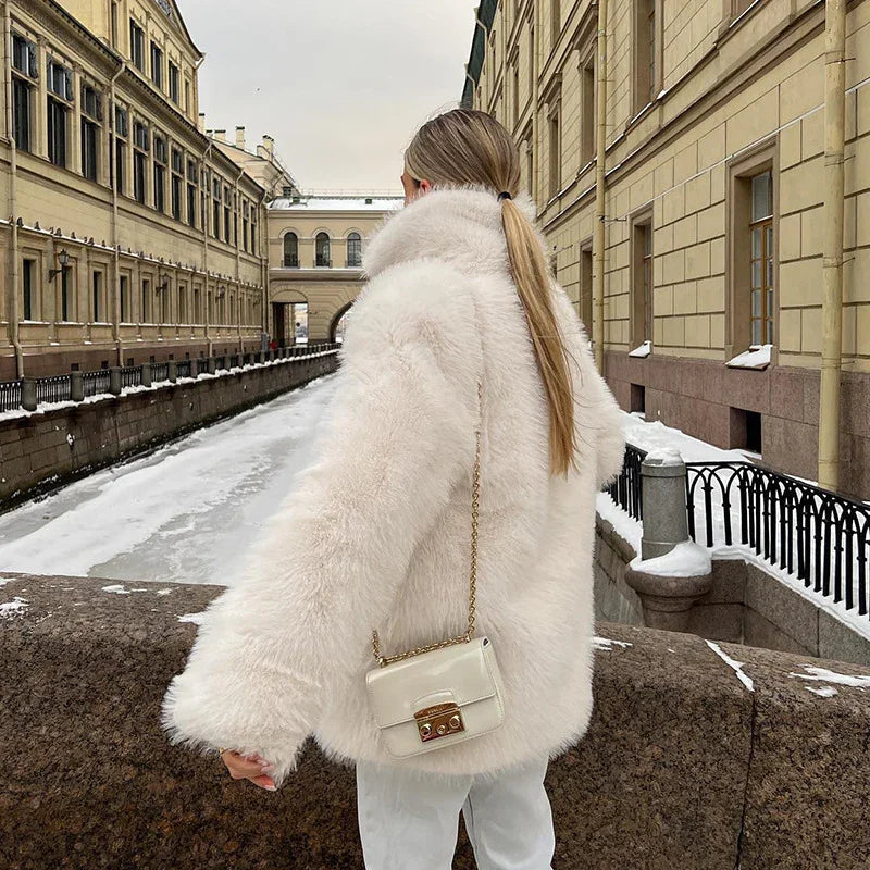 Woman in white faux fur coat and cream handbag standing by snowy canal and historic buildings