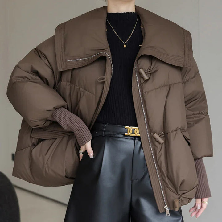 Brown oversized puffer jacket with wide collar, black ribbed sweater, and black leather pants with gold buckle belt
