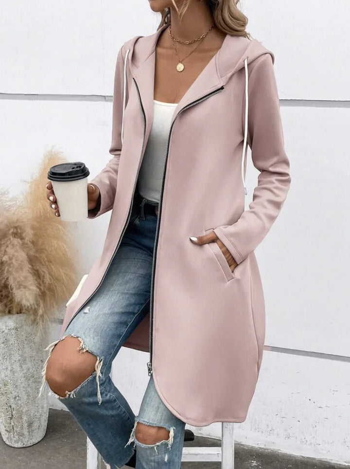 Woman wearing light pink zip-up hooded coat with white drawstrings, ripped blue jeans, and white top holding coffee cup