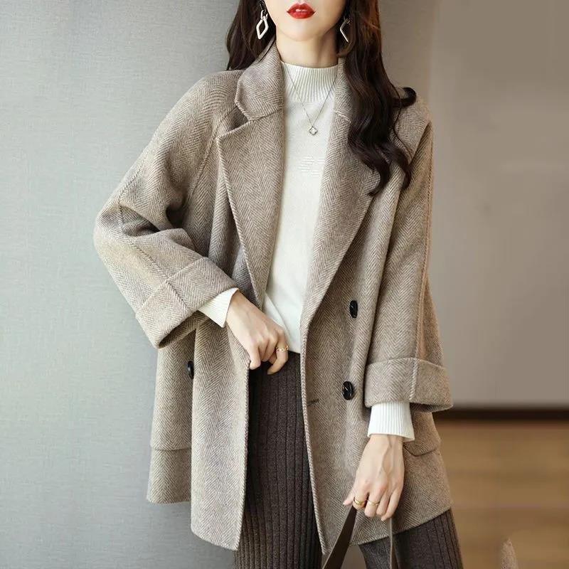 Woman wearing beige herringbone wool coat, white turtleneck sweater, and brown ribbed pants indoors