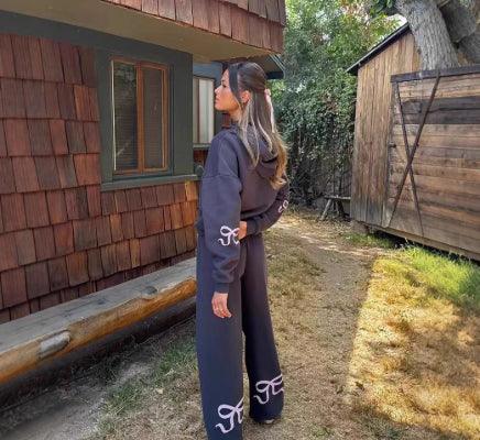 Woman wearing a black hoodie and wide-leg pants with white designs, standing beside a rustic wooden building