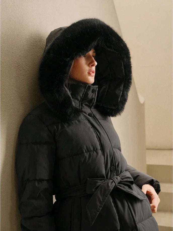 Woman wearing black puffer coat with fur-lined hood and belted waist, leaning against beige wall