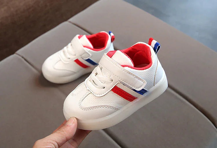 White toddler sneakers with red and blue stripes and red interior lining on a beige surface