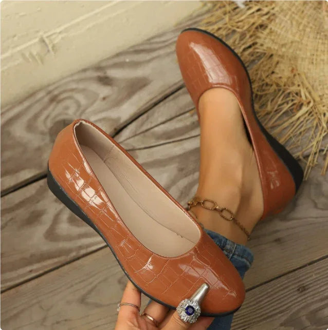 Brown crocodile pattern leather flats with black soles worn on wooden floor, featuring gold anklet and dark blue ring