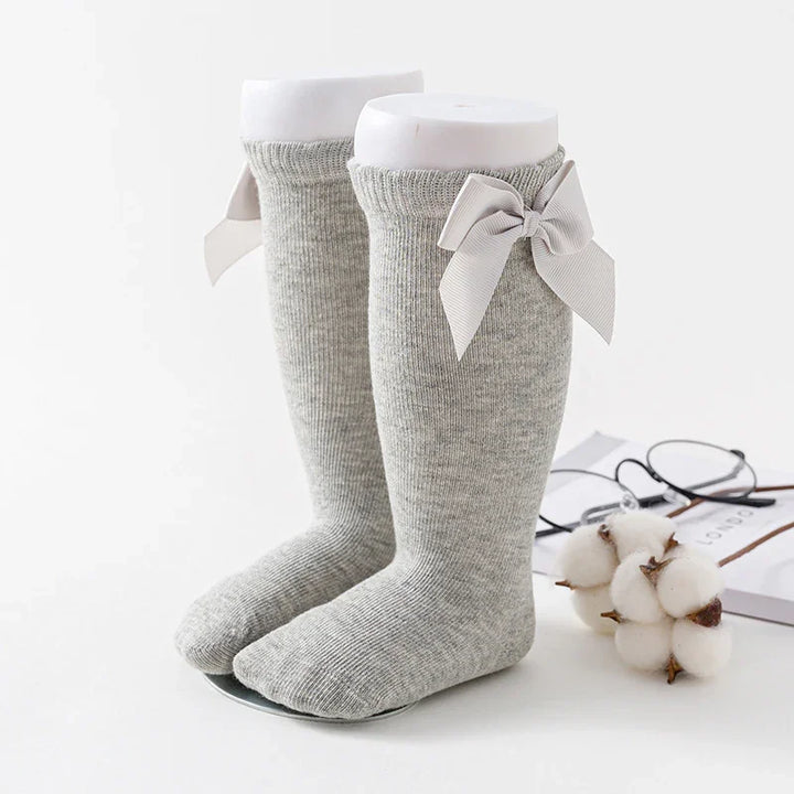 Gray knee-high socks with decorative bows on a white display stand beside glasses and cotton flowers