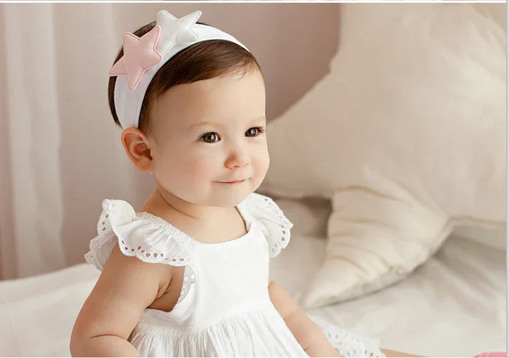 Smiling baby girl wearing white dress and headband with pink and white stars sitting on bed