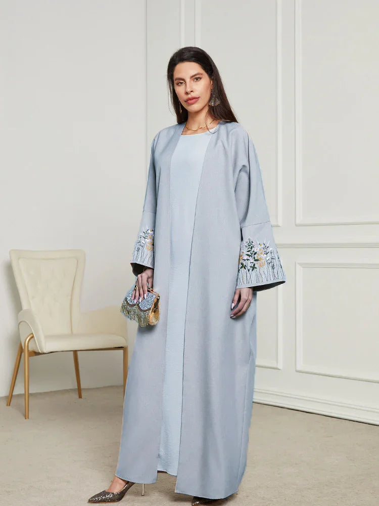 Woman wearing light blue abaya with floral embroidery on sleeves, standing in elegant room with beige chair
