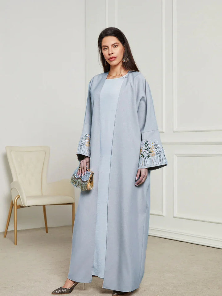 Woman wearing light blue abaya with floral embroidery on sleeves, standing in elegant room with beige chair