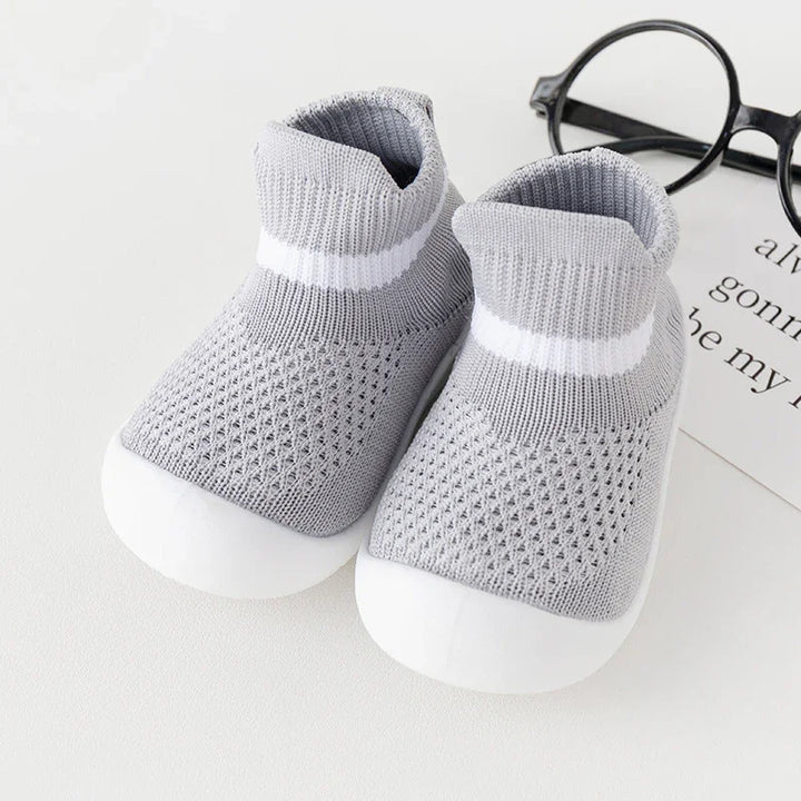 Pair of gray knit baby shoes with white soles and stripe, placed on white surface with glasses and paper