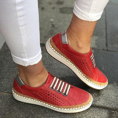 Red perforated casual sneakers with striped elastic bands and white soles worn with white pants on pavement