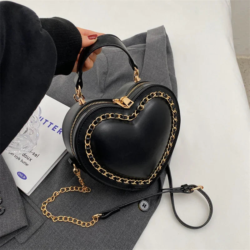 Black heart-shaped handbag with gold chain detail on gray fabric background