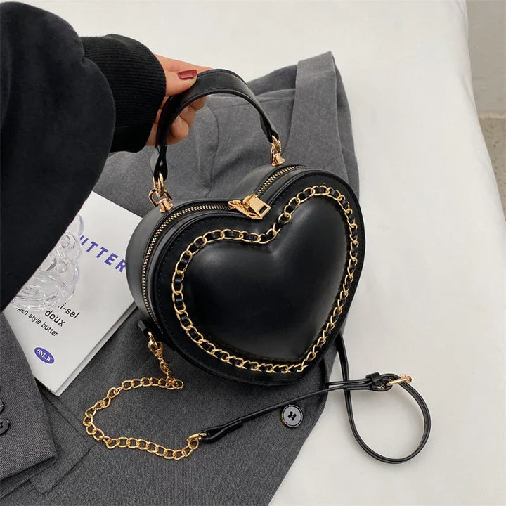 Black heart-shaped handbag with gold chain detail on gray fabric background