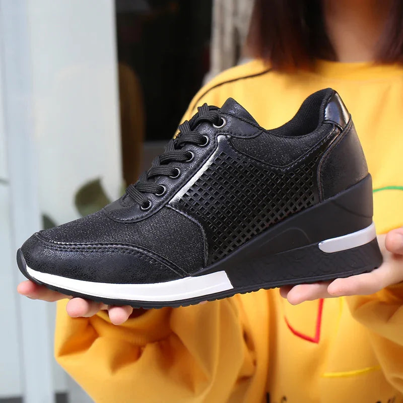 Black women's wedge sneakers with mesh pattern and white sole held by person in yellow sweatshirt