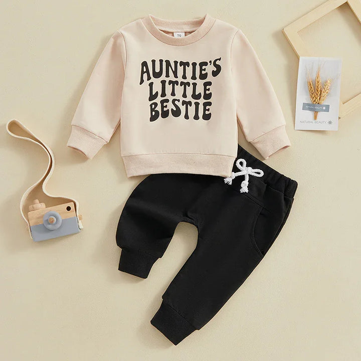 Beige toddler sweatshirt with 'Auntie's Little Bestie' text and black pants with white drawstring, flat lay