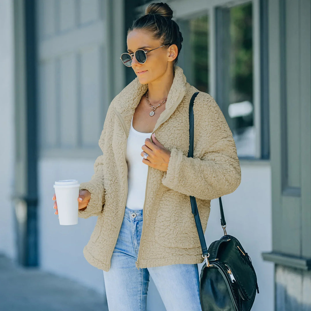 Woman wearing beige sherpa jacket, white top, light jeans, sunglasses, and carrying black bag outdoors