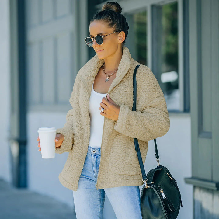 Woman wearing beige sherpa jacket, white top, light jeans, sunglasses, and carrying black bag outdoors