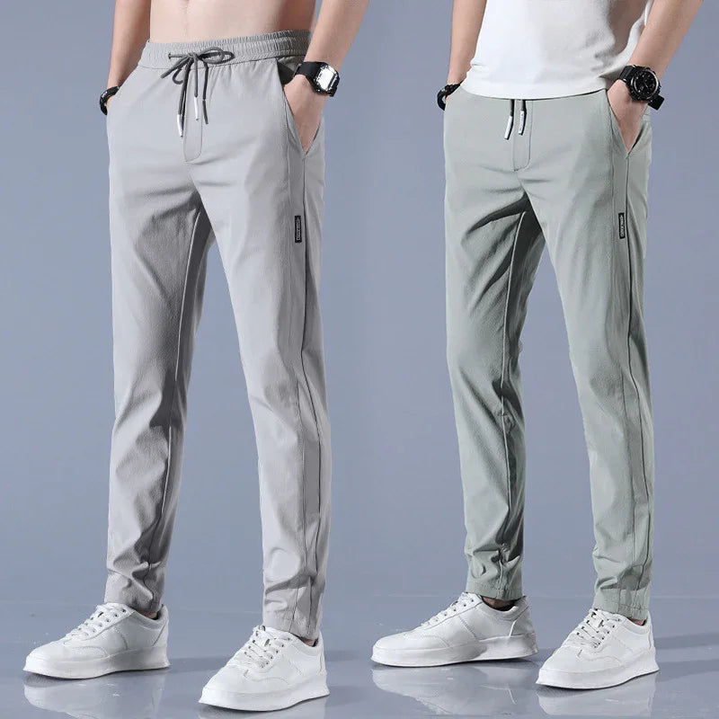 Two men wearing light gray and muted green slim fit casual jogger pants with white sneakers against a gray background