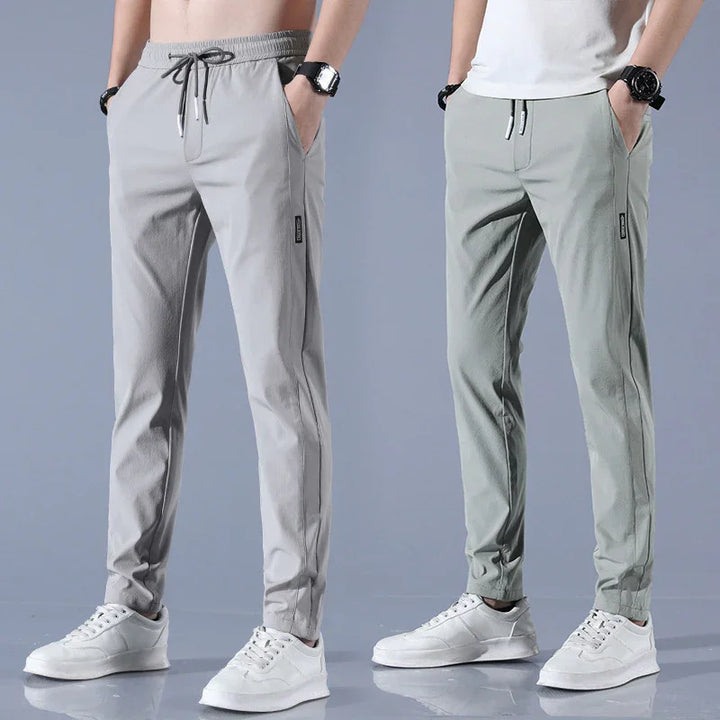 Two men wearing light gray and muted green slim fit casual jogger pants with white sneakers against a gray background