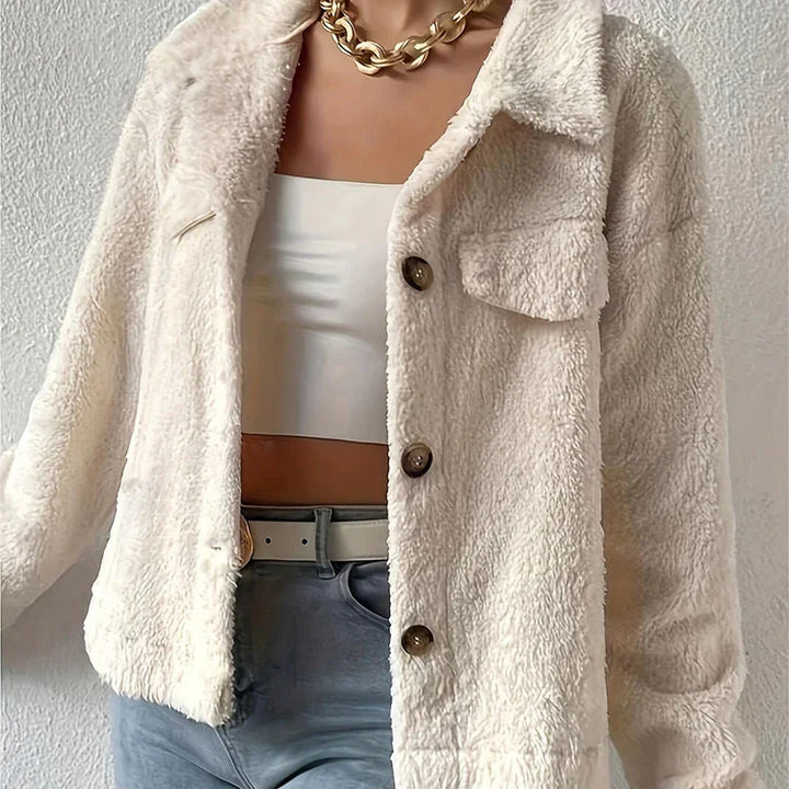 Woman wearing a cream sherpa jacket with buttons, white crop top, blue jeans, and chunky gold chain necklace
