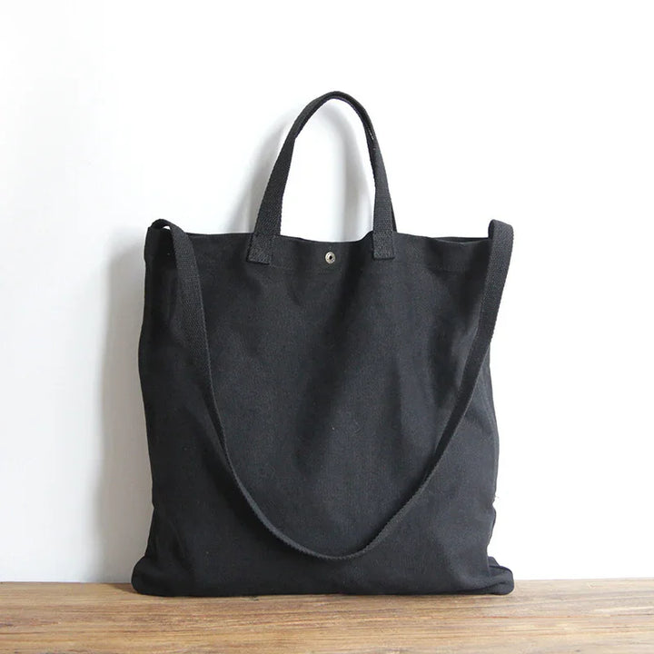 Black canvas tote bag with short handles and a long shoulder strap on wooden surface
