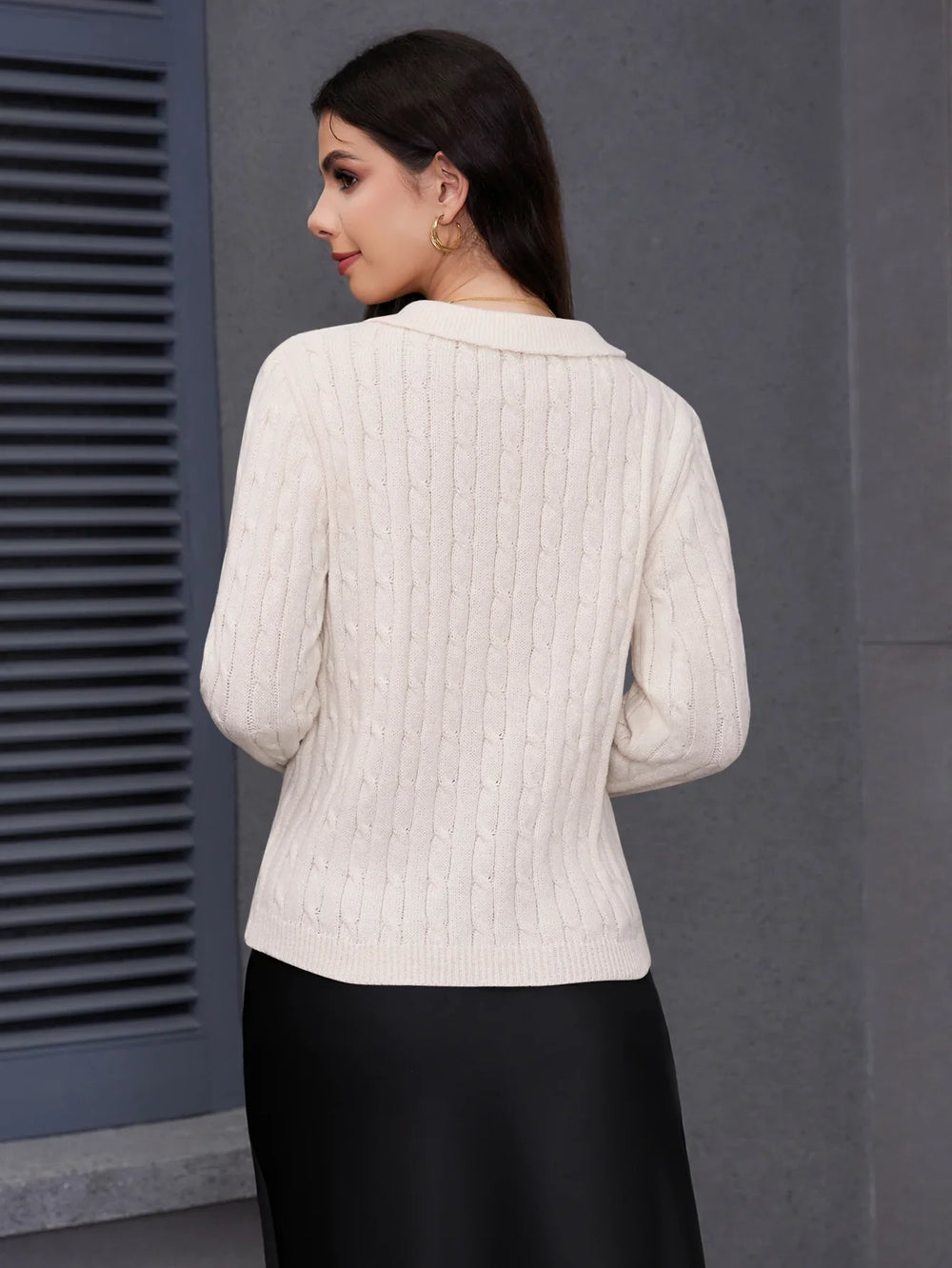 Woman wearing cream cable knit sweater and black skirt against gray wall background