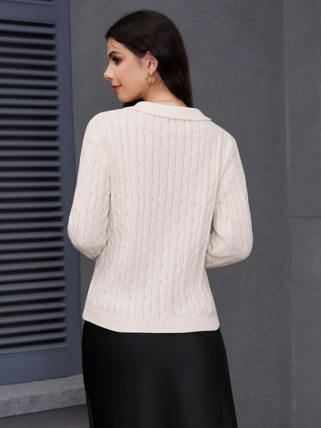 Woman wearing cream cable knit sweater and black skirt against gray wall background