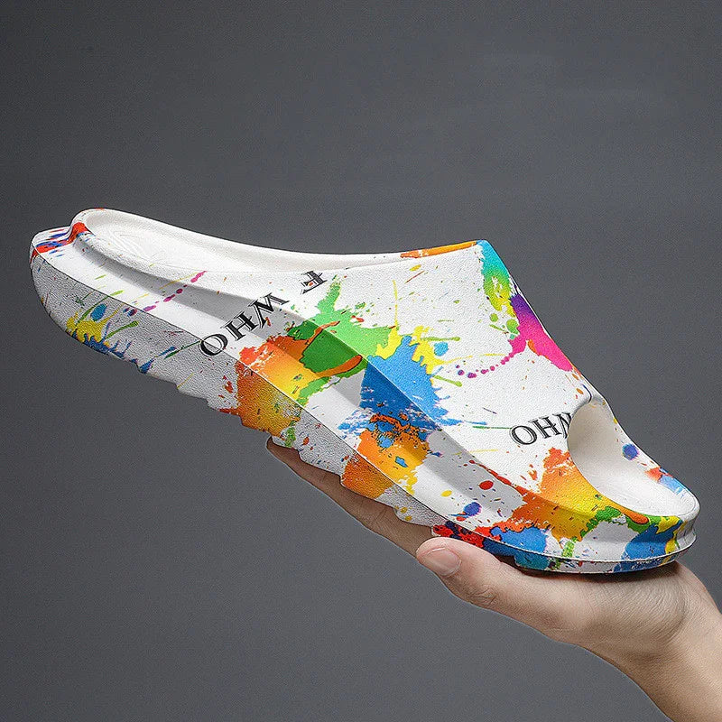 White slip-on sandal with colorful paint splatter design and black text held by hand on gray background