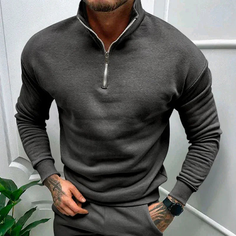 Man wearing dark gray half-zip sweatshirt with matching pants, tattoos on hands, and black wristwatch