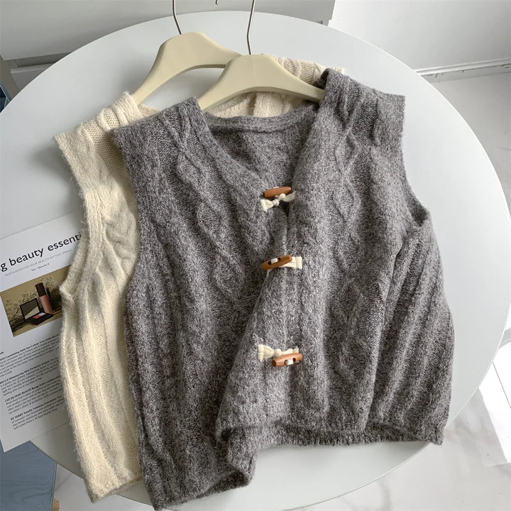 Two cable knit sleeveless vests on hangers, one gray with toggle buttons, one cream, on a white round table