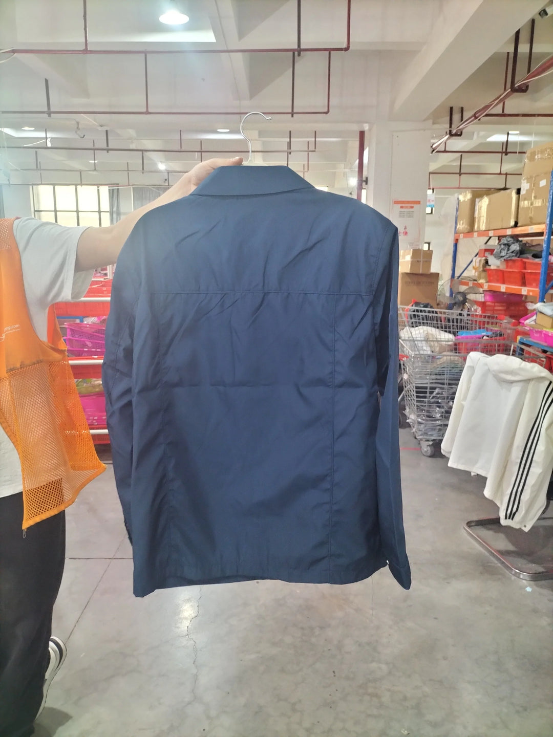 Person holding navy blue casual jacket on hanger inside warehouse with shelves and clothes