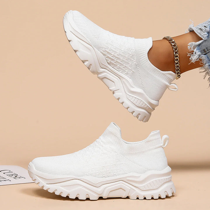 Pair of white slip-on knit sneakers with chunky sole, worn with ripped jeans and gold anklet