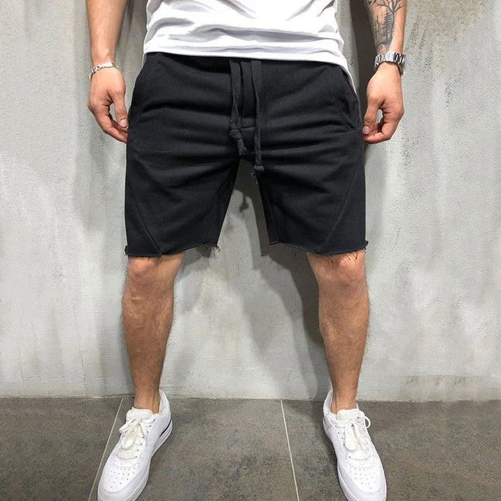Man wearing black drawstring shorts and white sneakers standing against a gray wall