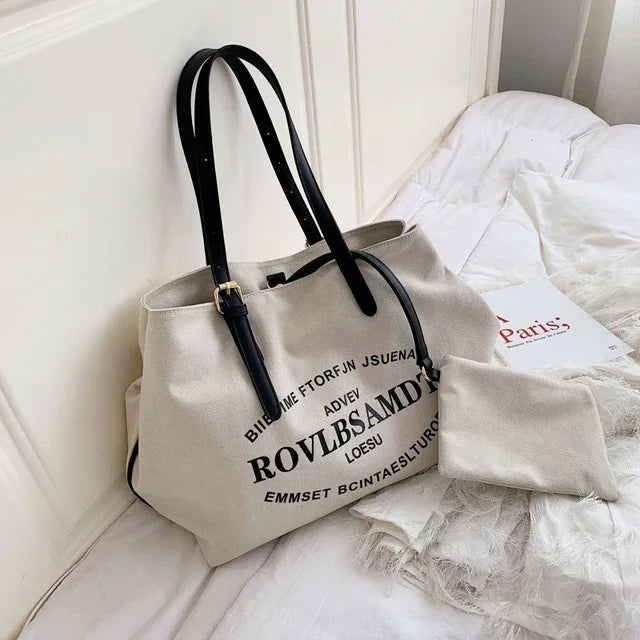 Beige canvas tote bag with black straps and bold black text on white bed with matching pouch and Paris book