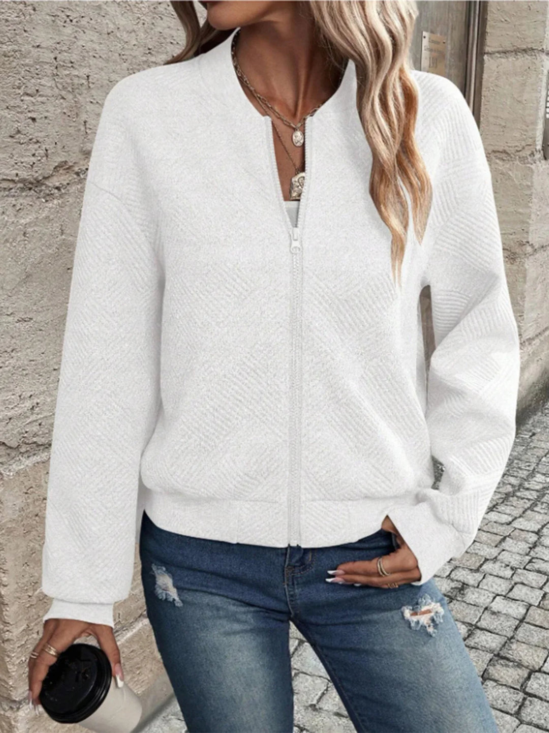 Woman wearing white textured zip-up jacket and ripped blue jeans, holding a coffee cup on cobblestone street