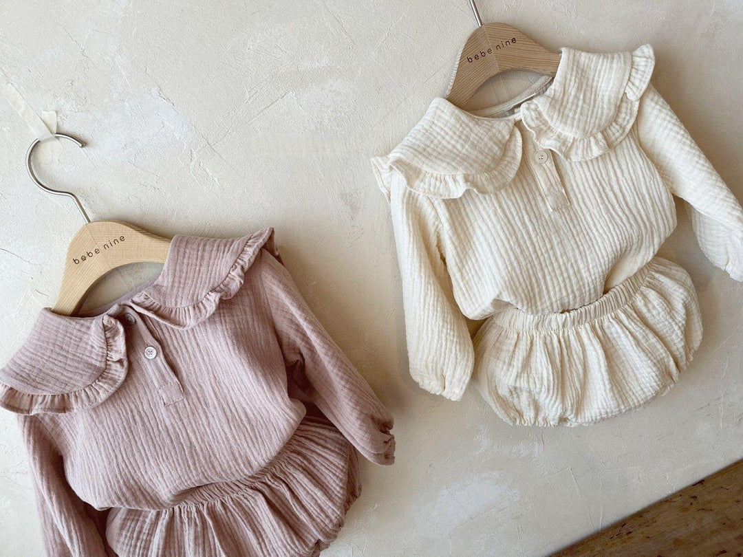 Two long-sleeve textured baby outfits with ruffled collars in soft beige and cream on wooden hangers