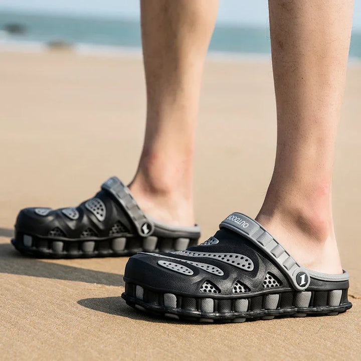 Black and gray outdoor slide sandals with ventilation holes worn on feet standing on sandy beach