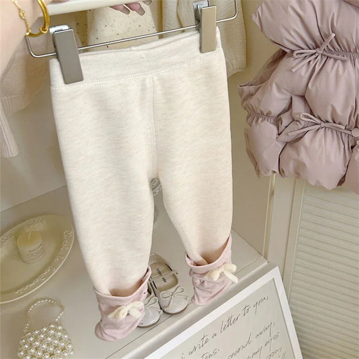 Cream toddler leggings with pink rolled cuffs and bow detail hanging on hanger in a cozy nursery room