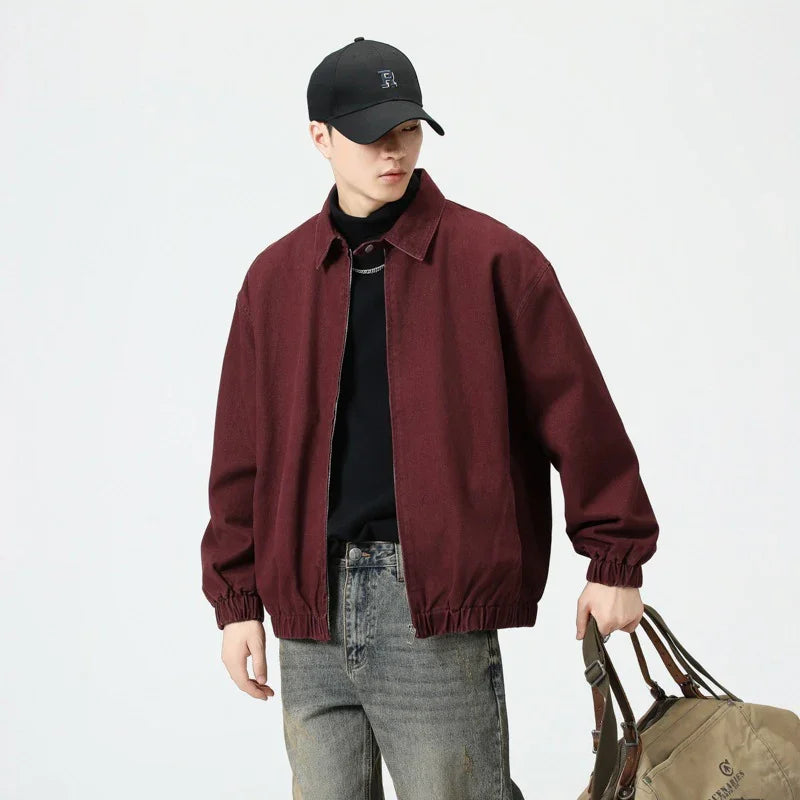 Man wearing burgundy bomber jacket, black cap, black shirt, gray jeans, holding brown duffel bag